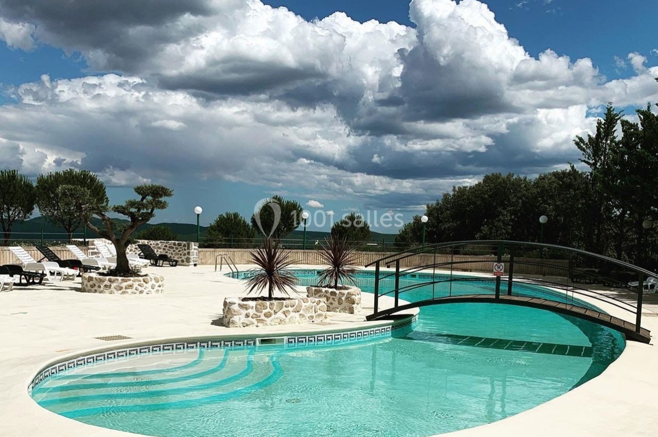 Piscine extérieure avec pont en arc, entourée de chaises longues, végétation et ciel nuageux en arrière-plan.