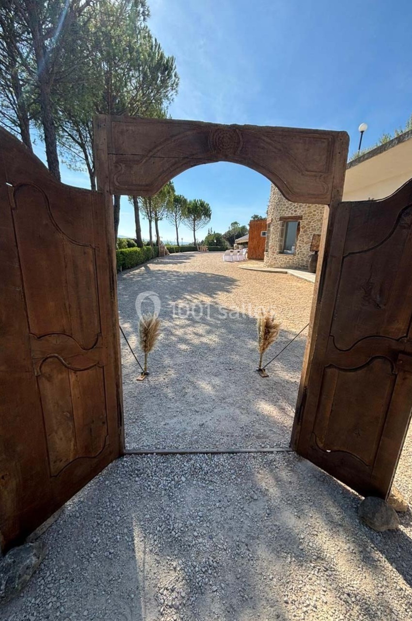 Portail en bois ouvert sur une allée gravillonnée, bordée d'arbres et menant à un bâtiment en pierre sous un ciel dégagé.