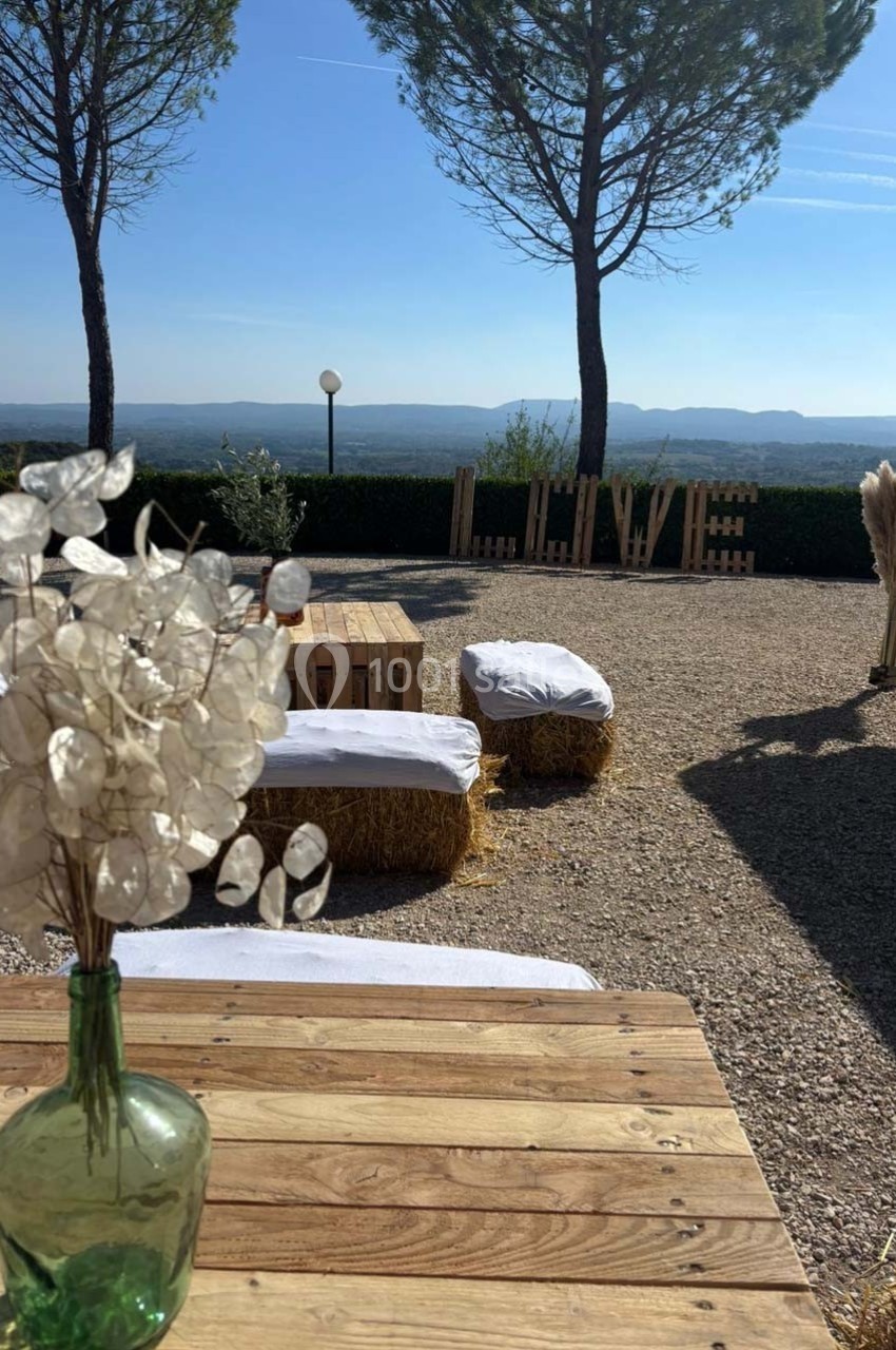 Espace extérieur avec bancs en bottes de foin, table en bois, vase vert et vue dégagée sur un paysage vallonné.