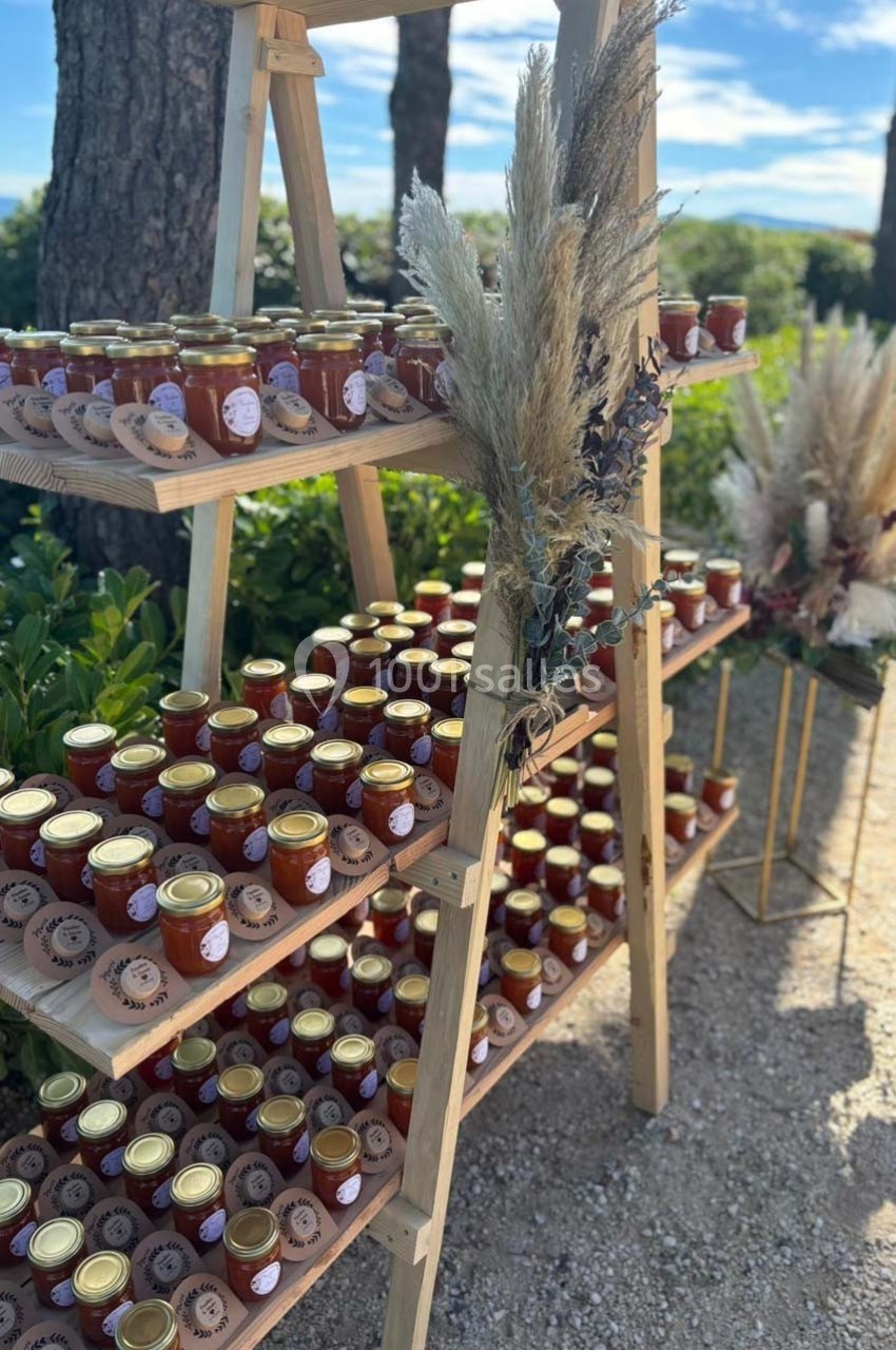 Étagère en bois remplie de pots de confiture artisanale, décorée de fleurs séchées, en extérieur sous un ciel dégagé.