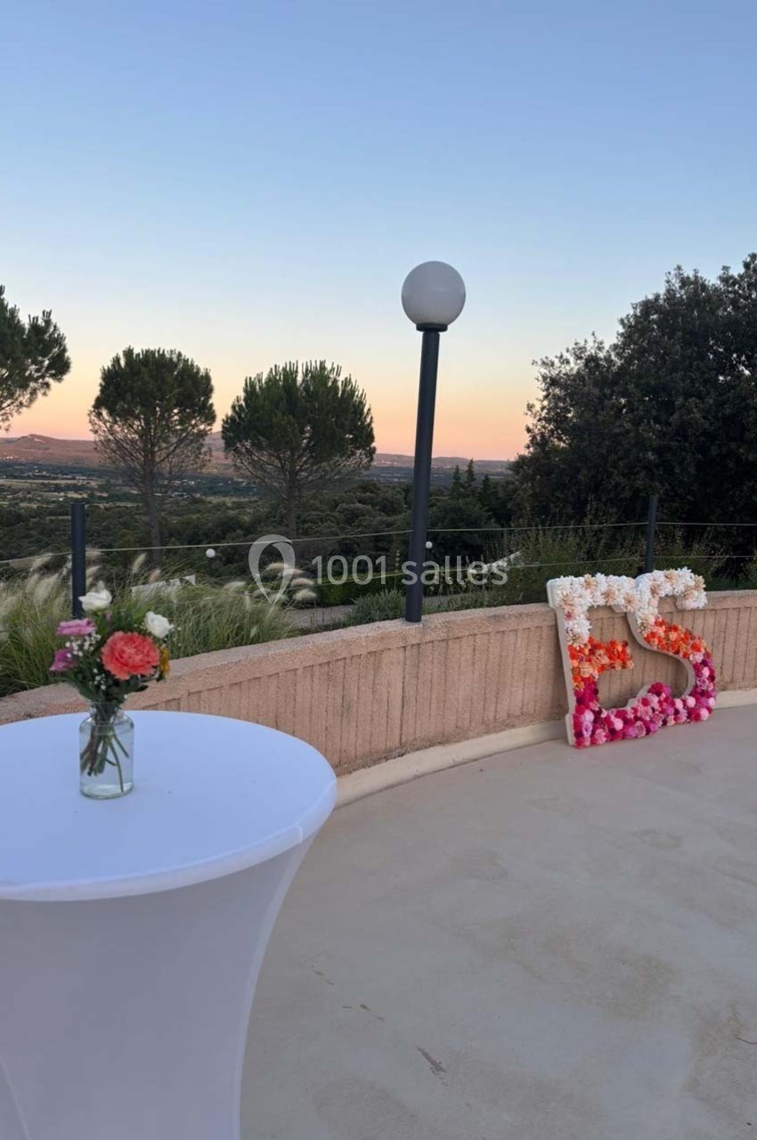 Terrasse extérieure au coucher du soleil avec table haute décorée de fleurs et initiales en fleurs près d'une rambarde.