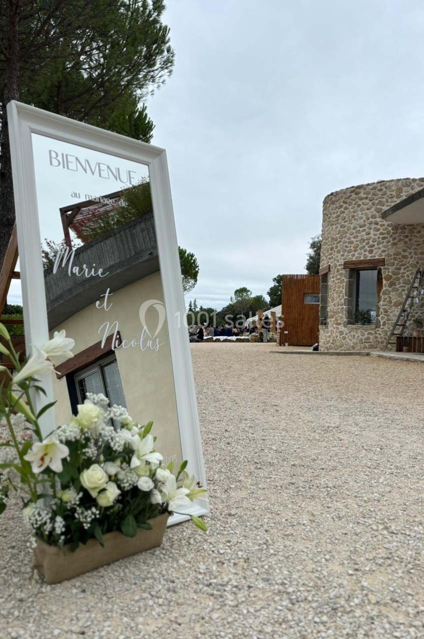 Panneau de bienvenue avec des fleurs blanches devant un bâtiment en pierre dans un cadre extérieur.