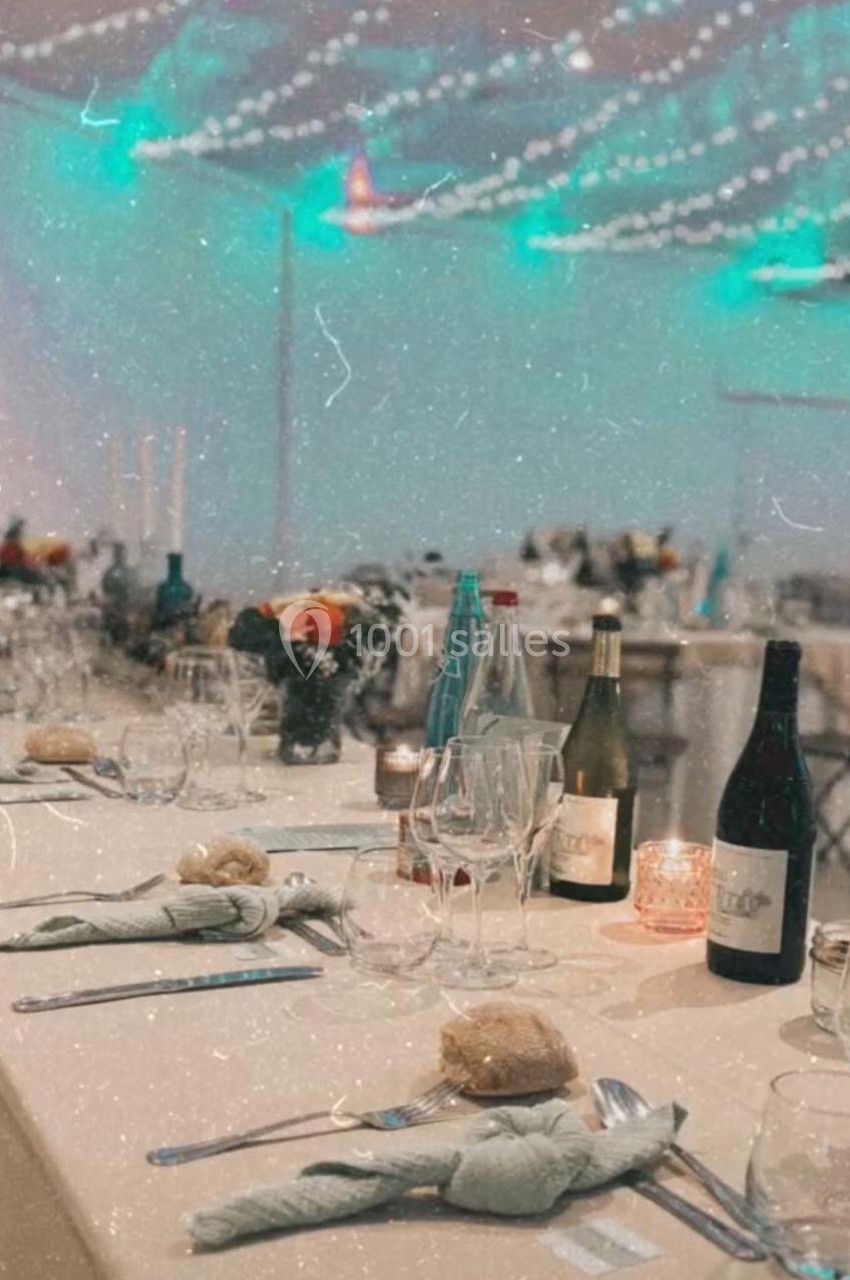 Table dressée pour un repas festif avec nappes blanches, verres, bouteilles de vin et décorations lumineuses en arrière-plan.