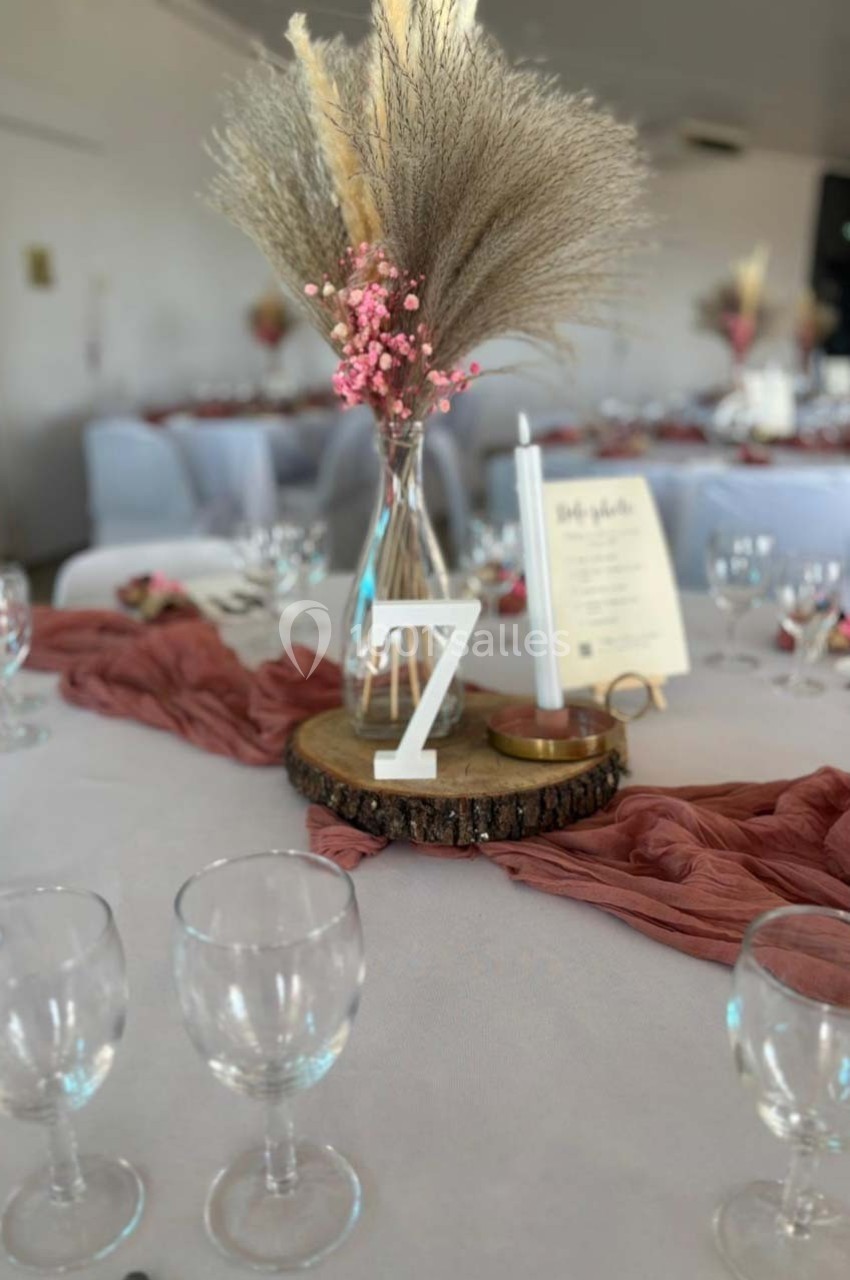 Centre de table avec vase de fleurs séchées, chiffre 7, bougie et menu sur une base en bois, nappes blanches.