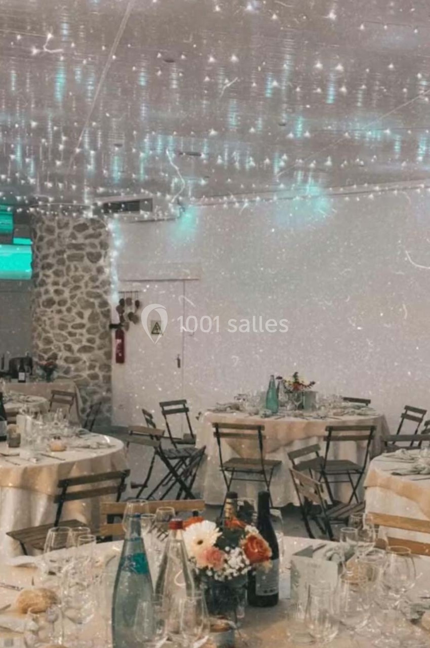 Salle de réception décorée avec des guirlandes lumineuses, tables rondes dressées et bouquets de fleurs.