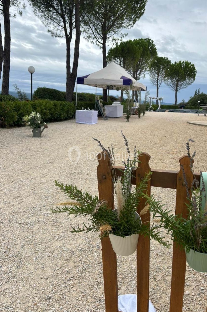 Allée extérieure gravillonnée bordée de végétation, avec une tente blanche et des décorations florales suspendues.