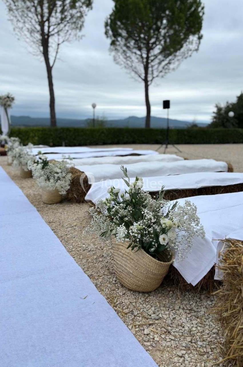 Allée extérieure décorée pour une cérémonie, avec bancs en bottes de paille recouverts de tissu blanc et fleurs en paniers.