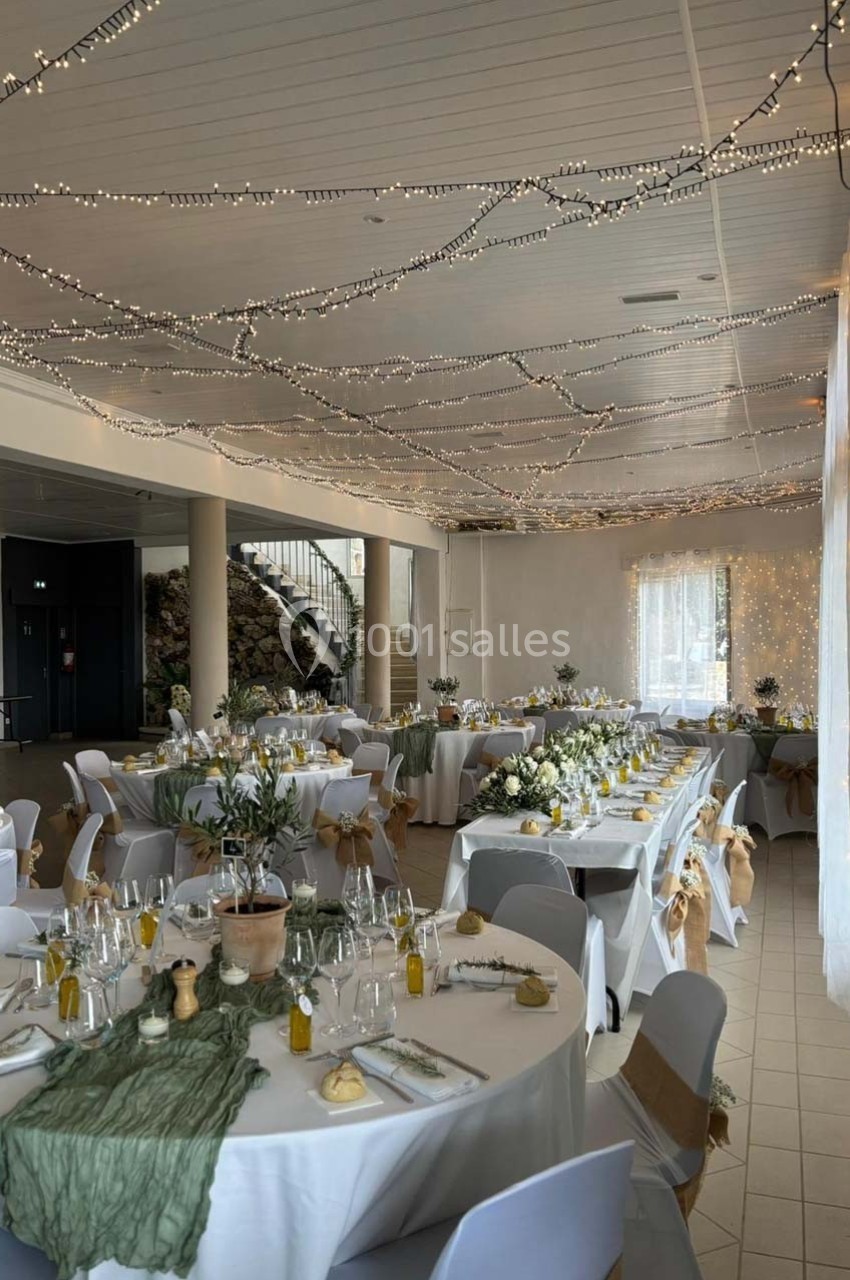 Salle de réception décorée avec des guirlandes lumineuses, tables dressées avec nappes blanches et chemins de table verts.