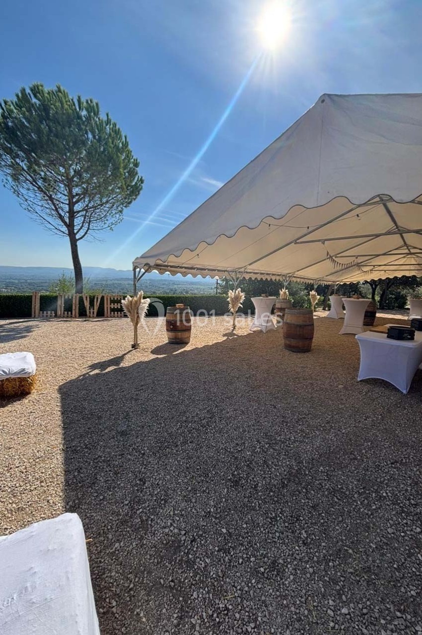 Tente blanche installée en extérieur sur un sol gravillonné, avec des tables décorées et un paysage ensoleillé en arrière…