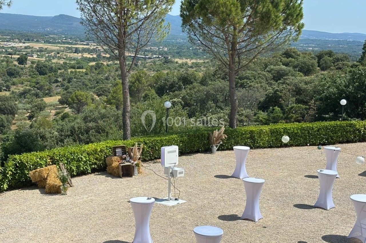 Espace extérieur avec tables hautes blanches, vue sur un paysage vallonné et arbres en arrière-plan.
