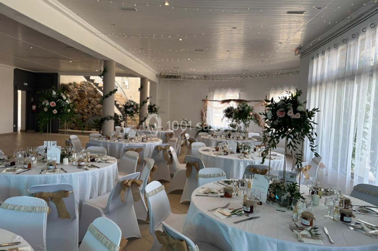 Salle de réception décorée pour un mariage, avec tables rondes dressées, chaises ornées de rubans et guirlandes lumineuses…