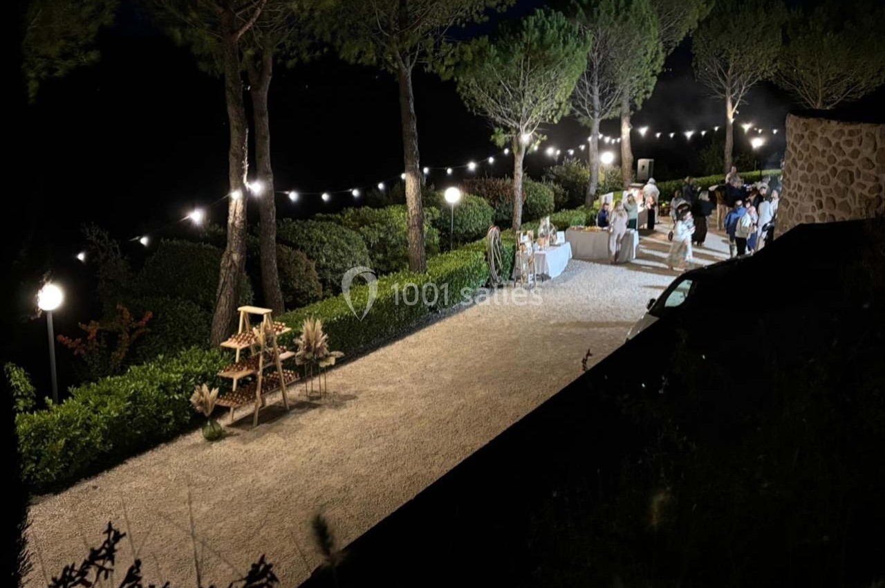 Allée éclairée de guirlandes lumineuses entourée d'arbres, avec des tables et des invités lors d'un événement en soirée.