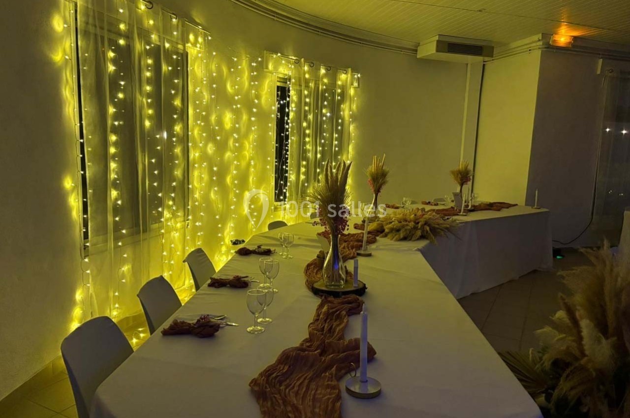 Salle décorée avec des guirlandes lumineuses, tables dressées avec nappes blanches, bougies et compositions florales.