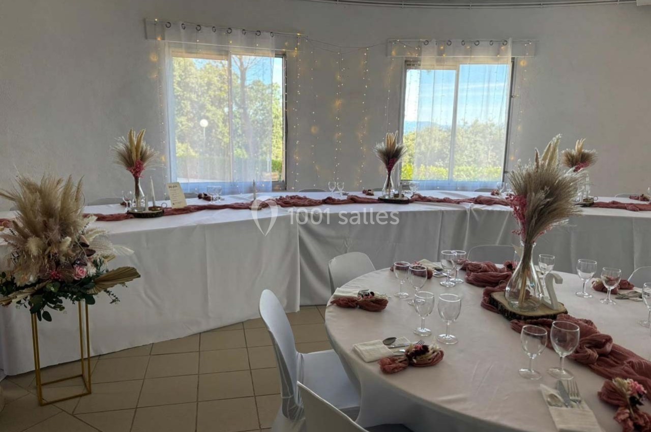 Salle décorée pour un événement avec tables rondes et nappes blanches, ornées de fleurs séchées et de vaisselle.