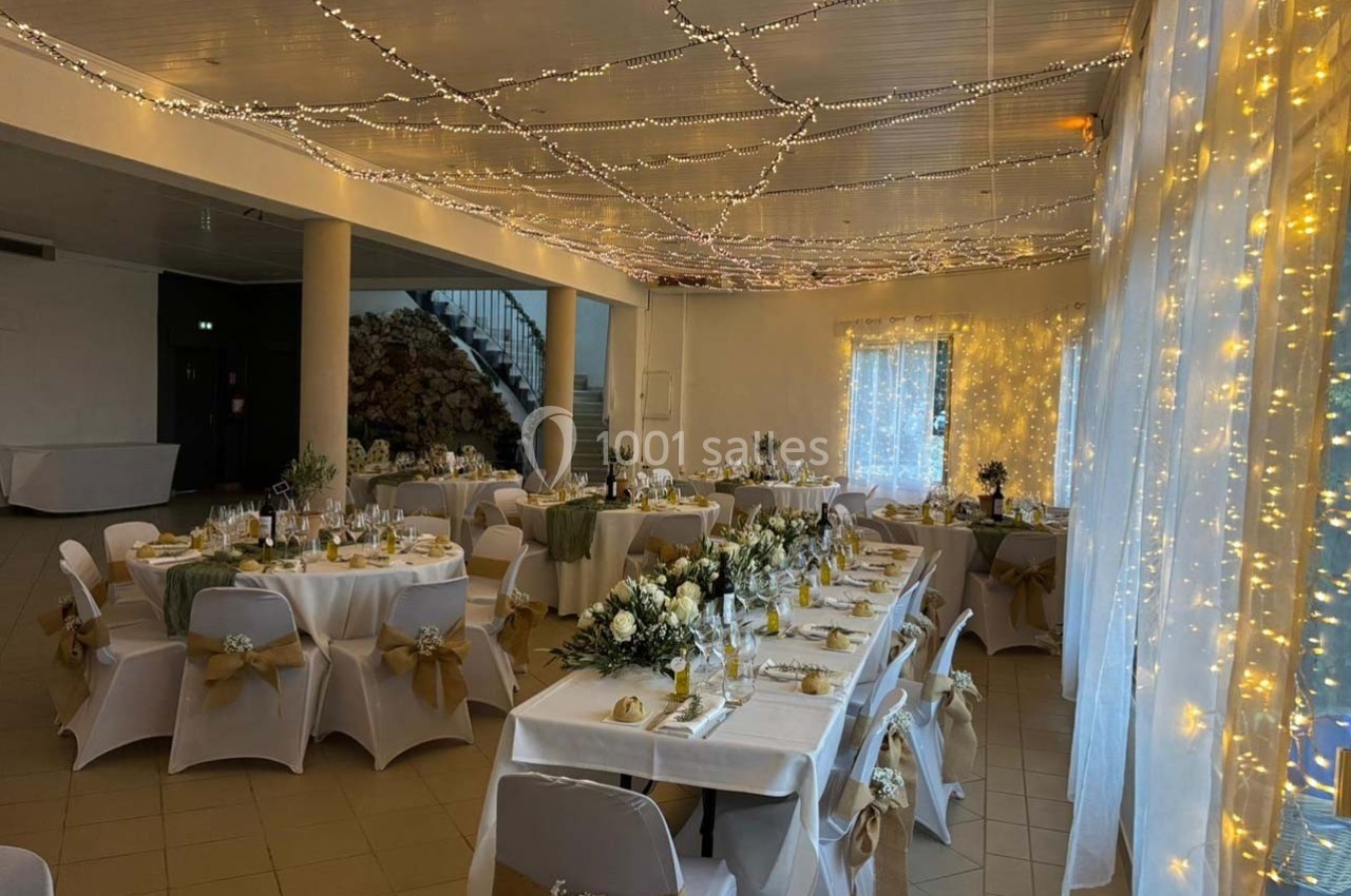 Salle de réception décorée pour un mariage, avec tables dressées, nappes blanches, fleurs et guirlandes lumineuses.
