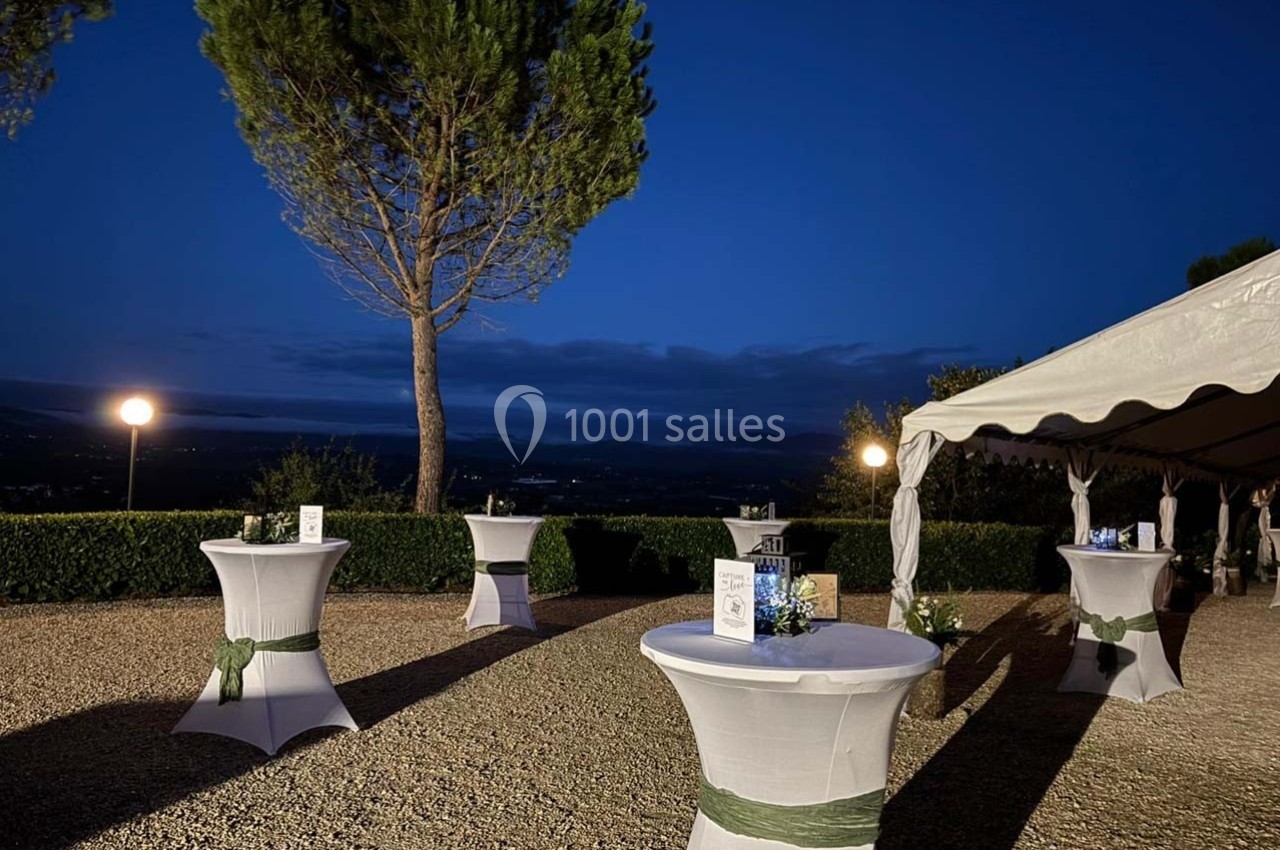 Tables hautes décorées sous un ciel nocturne, près d'un arbre et d'une tente éclairée dans un espace extérieur.