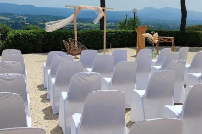 Chaises blanches alignées en extérieur face à une arche décorée, avec vue sur un paysage vallonné et des arbres.