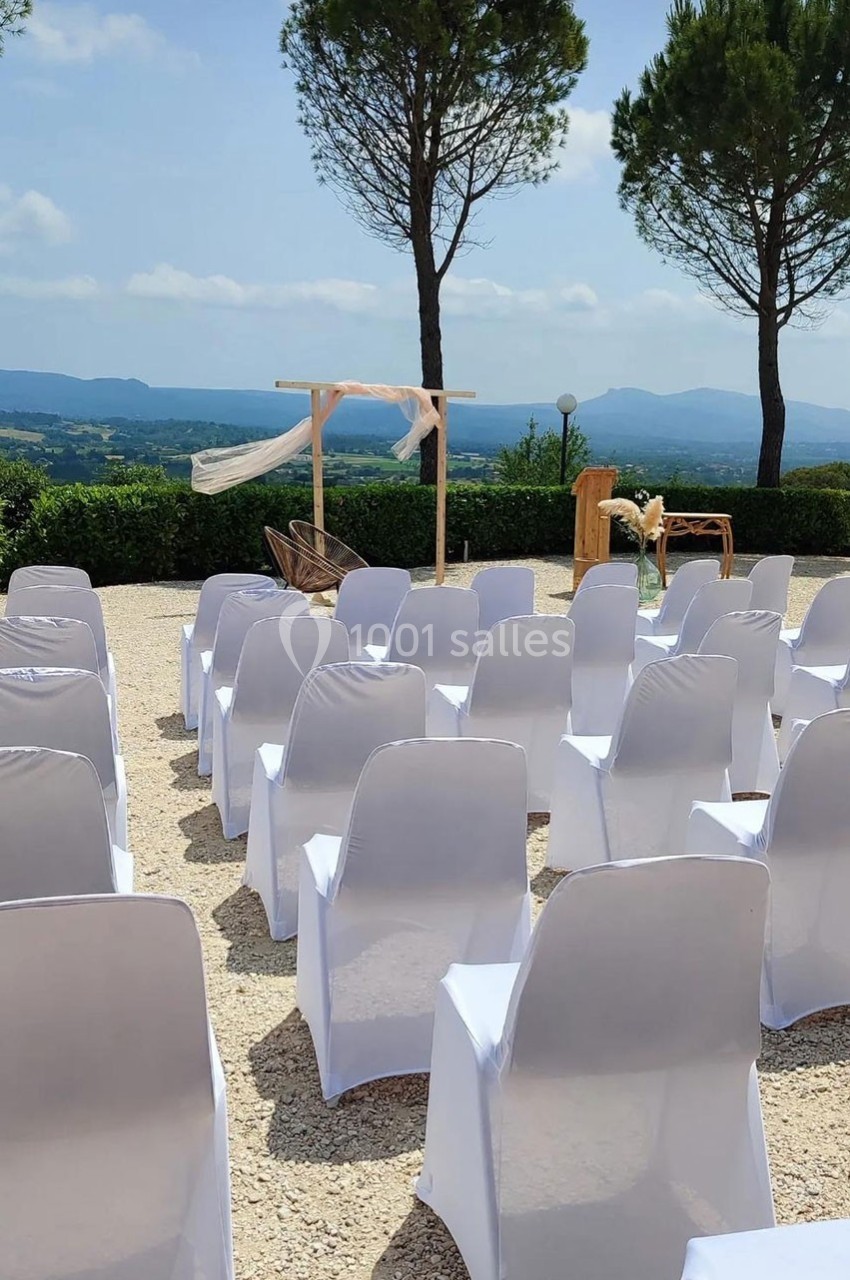Chaises blanches alignées en extérieur face à une arche décorée, avec vue sur un paysage vallonné et des arbres.