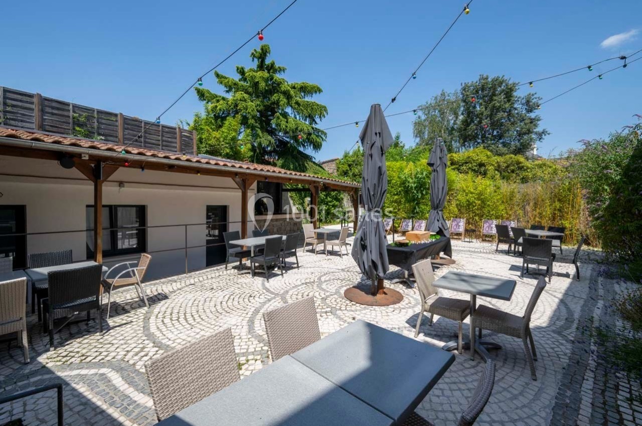 Terrasse extérieure pavée avec tables, chaises, parasols fermés et guirlandes lumineuses sous un ciel dégagé.