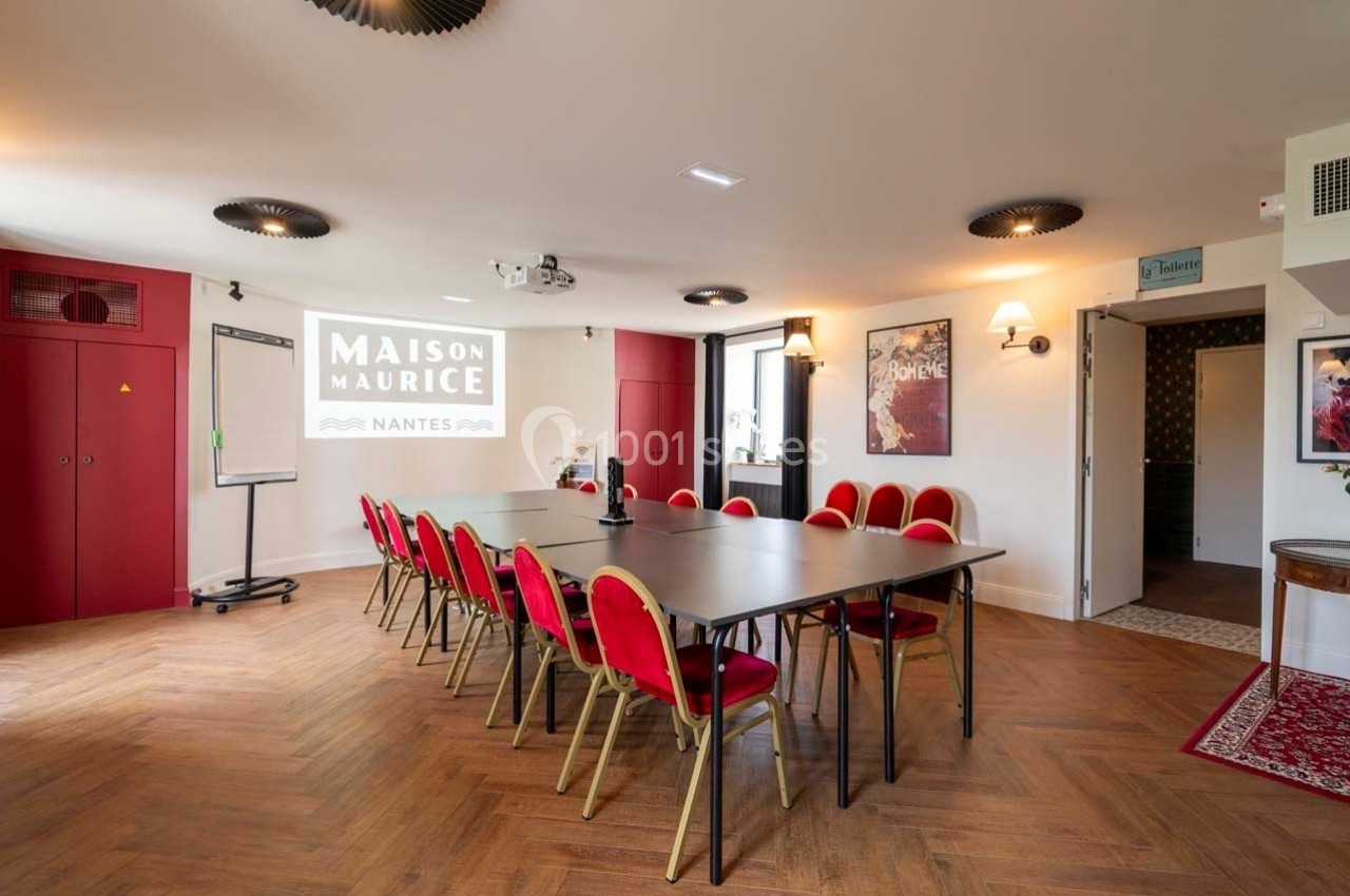 Salle de réunion lumineuse avec table rectangulaire, chaises rouges, projecteur et écran affichant ’Maison Maurice’.