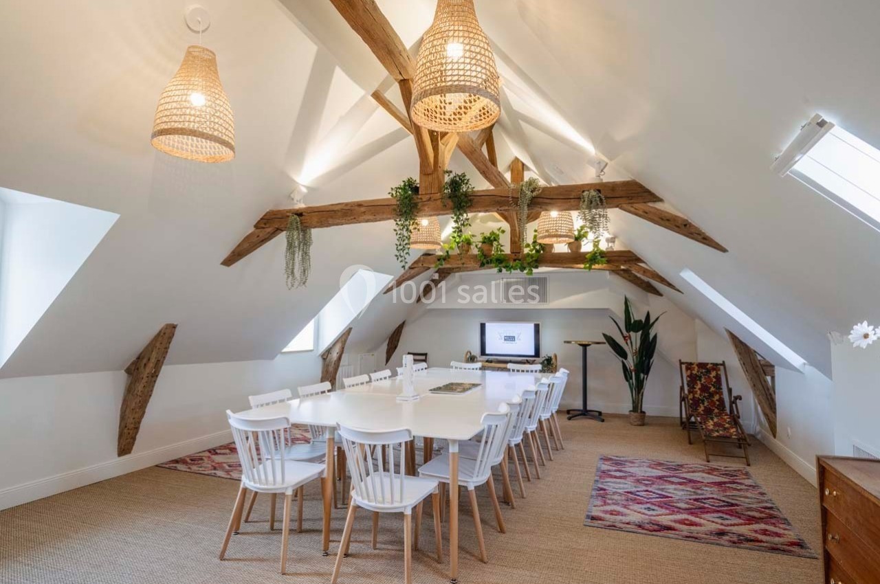 Salle de réunion lumineuse sous combles avec poutres apparentes, grande table blanche, chaises et décorations végétales.
