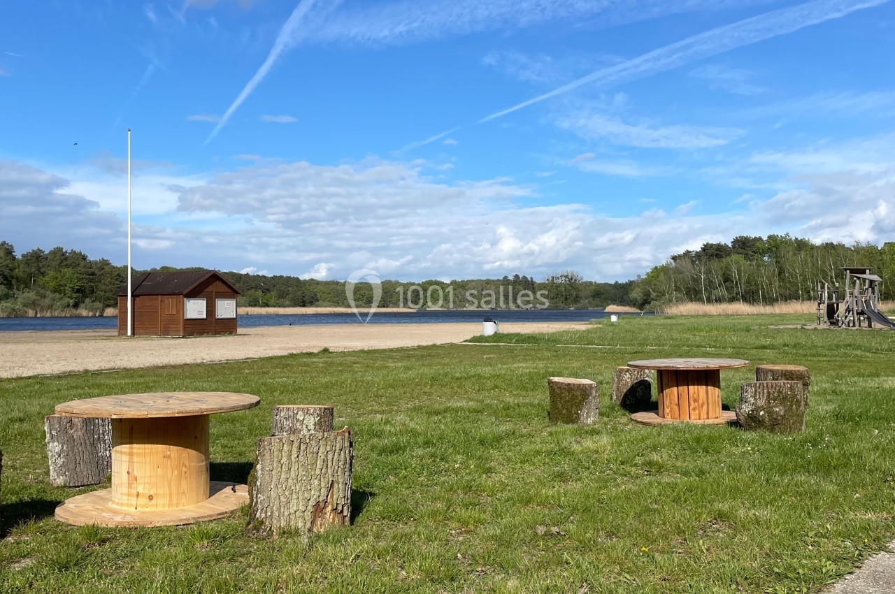 Tables et sièges en bois disposés sur une pelouse près d'un lac, avec une cabane et un ciel dégagé en arrière-plan.