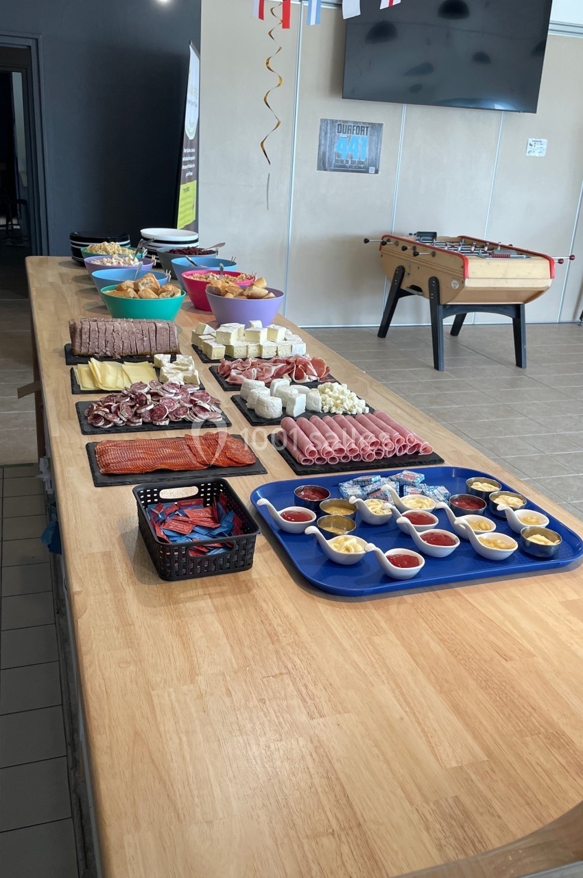 Table en bois garnie de divers plats, charcuteries, fromages, sauces et snacks, dans une salle avec baby-foot et décorations.