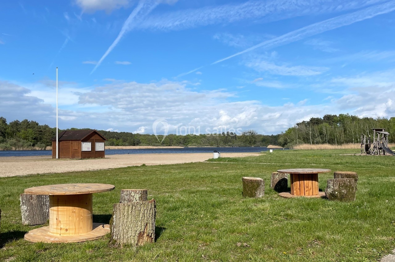 Tables et sièges en bois installés sur une pelouse près d'une plage avec un plan d'eau et un ciel dégagé en arrière-plan.