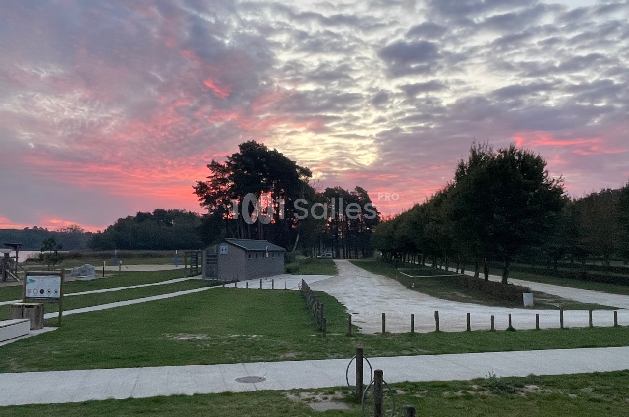 Coucher de soleil avec ciel nuageux coloré, arbres, chemins en gravier et petite construction dans un espace vert.