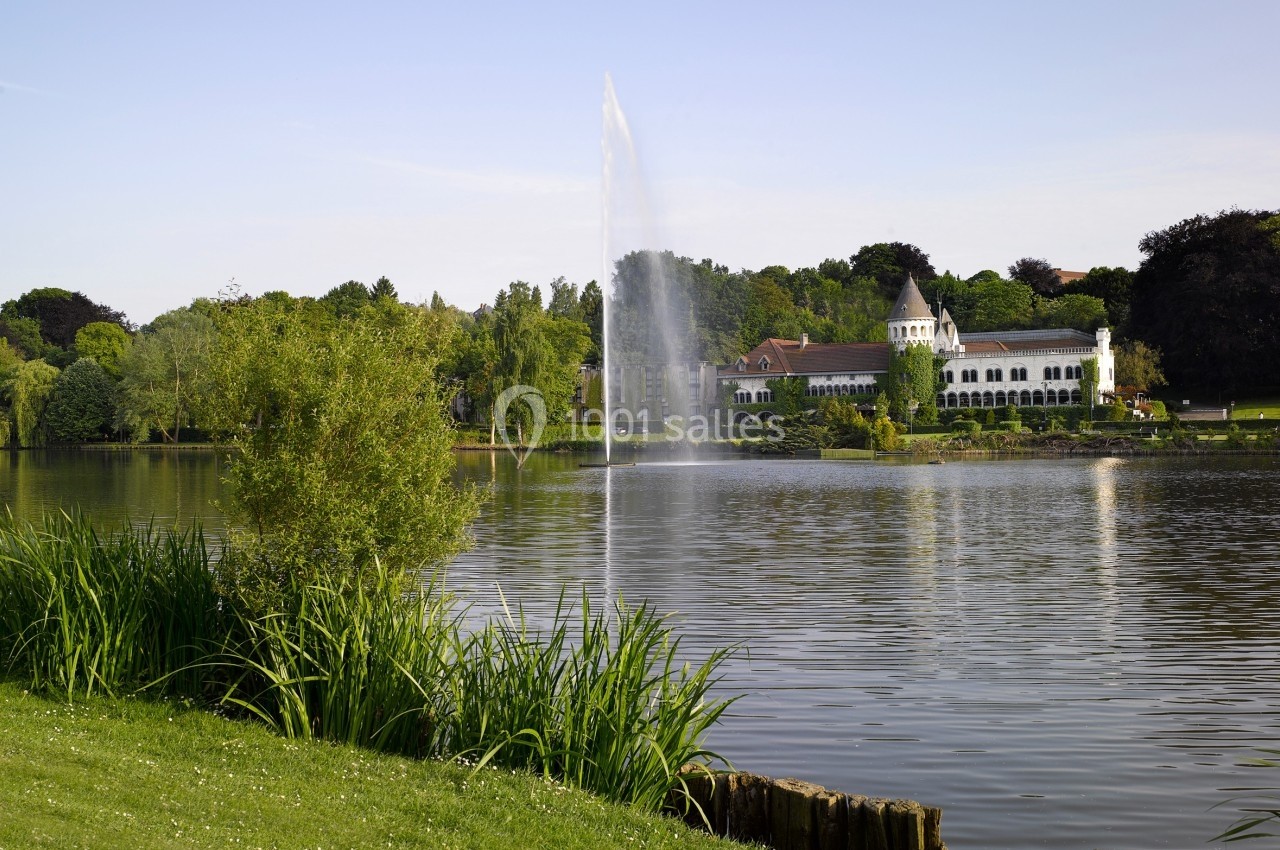 Vue d'un lac bordé de verdure avec une fontaine centrale et un bâtiment blanc en arrière-plan.