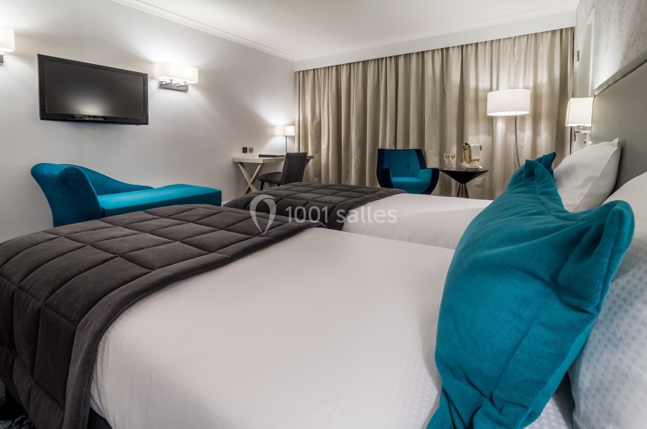 Chambre d'hôtel avec deux lits simples, fauteuils bleus, bureau, rideaux beiges et éclairage doux.