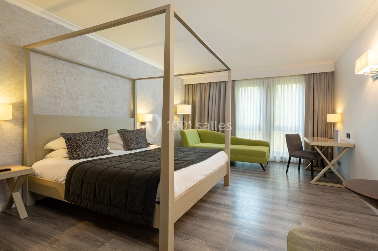 Chambre d'hôtel lumineuse avec lit à baldaquin, canapé vert, bureau et grande fenêtre avec rideaux.