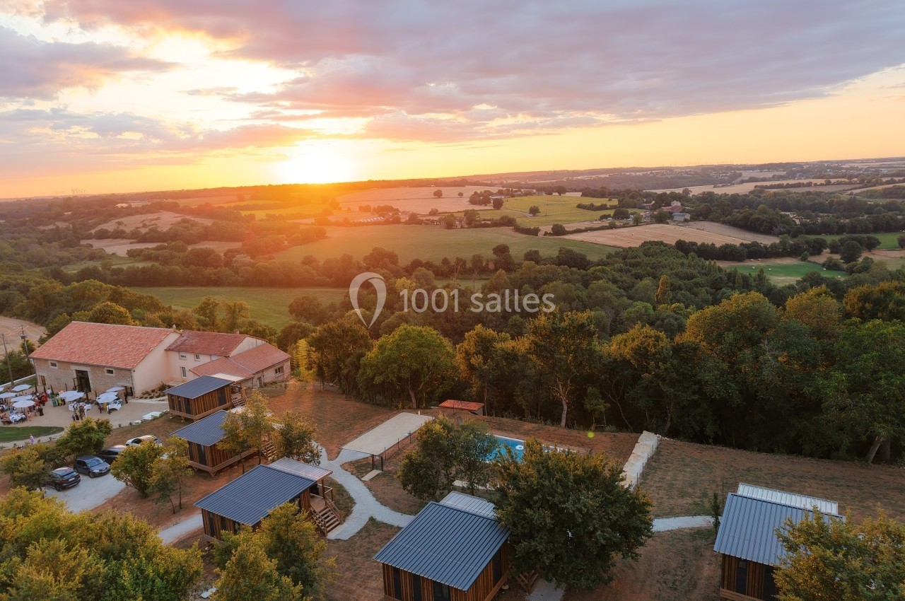 Vue aérienne d'un domaine rural au coucher du soleil avec chalets en bois, piscine et paysage vallonné.