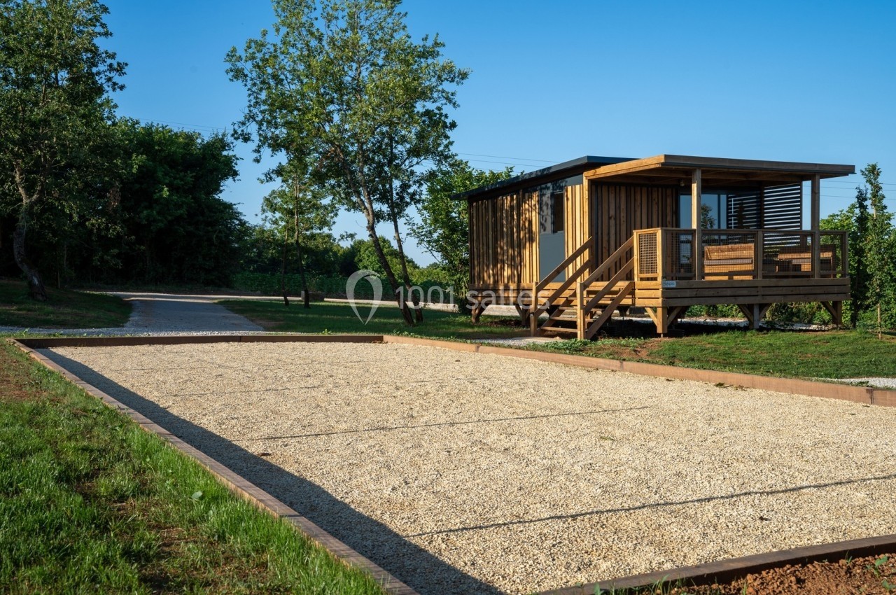 Terrain de pétanque en gravier devant un chalet en bois entouré de verdure, sous un ciel dégagé.