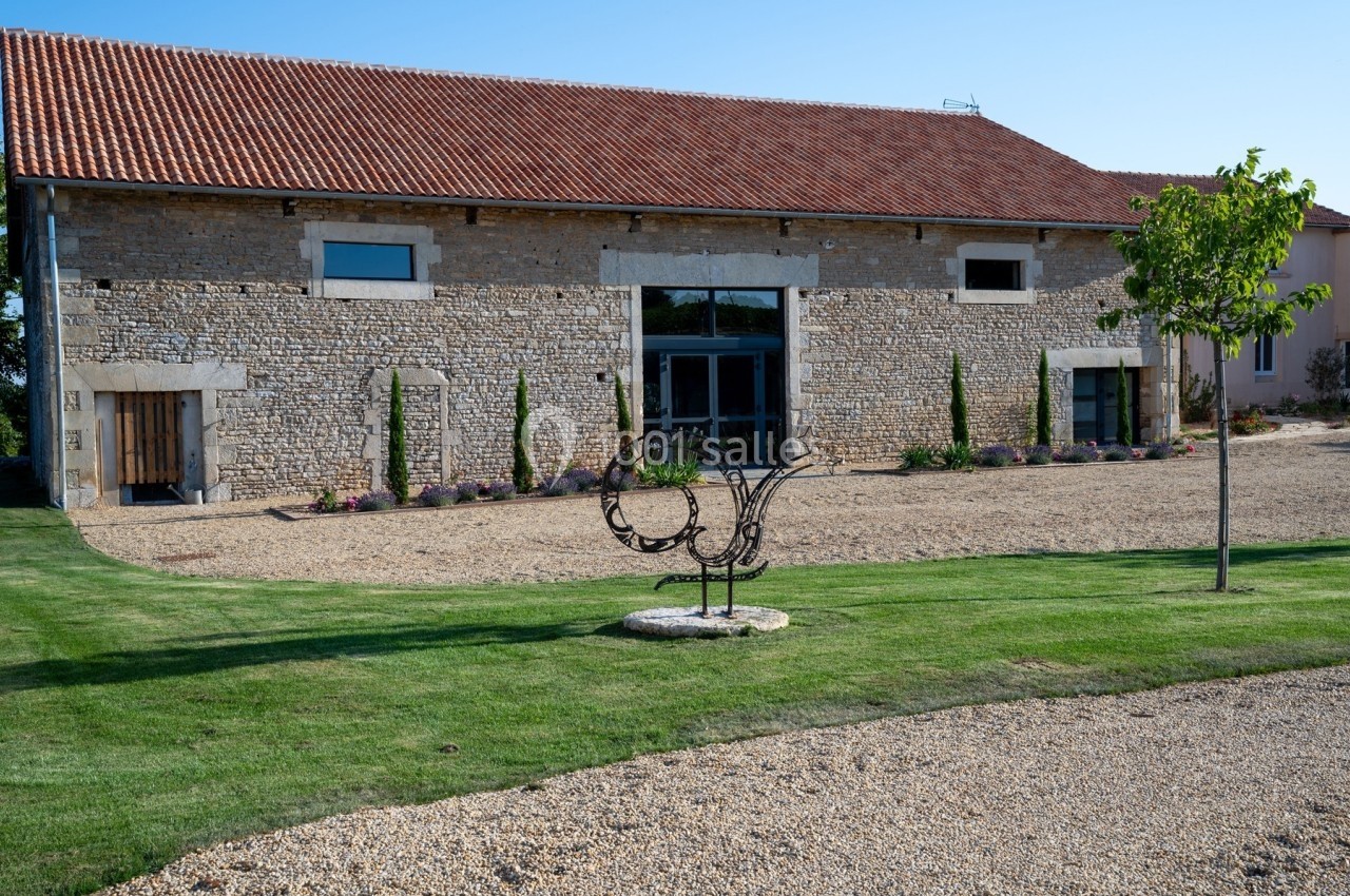 Bâtiment en pierre avec toit en tuiles rouges, entouré de pelouse, graviers et une sculpture métallique au premier plan.
