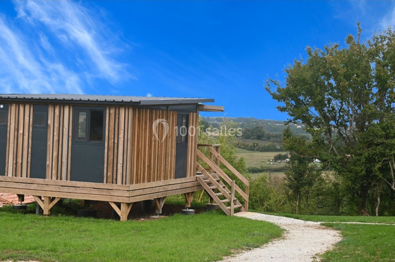 Cabane en bois surélevée avec terrasse, située dans un paysage rural verdoyant sous un ciel bleu.