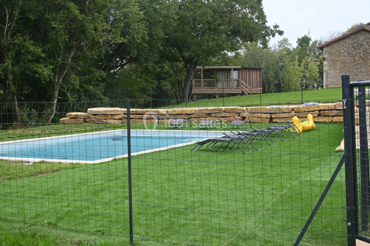Piscine extérieure entourée de chaises longues sur pelouse, avec clôture et cabane en bois en arrière-plan.