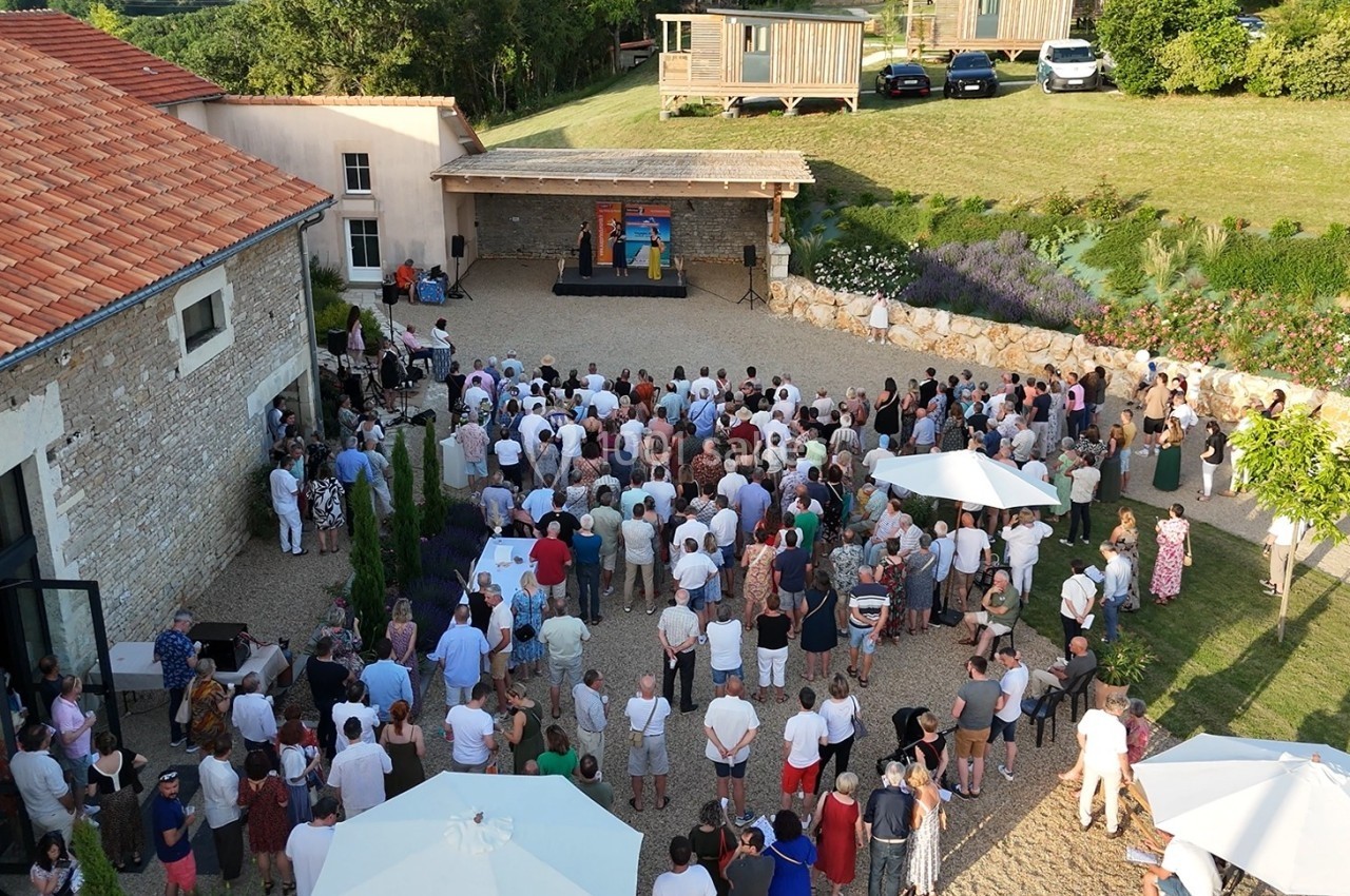 Vue aérienne d'une assemblée en plein air devant une scène dans une cour entourée de bâtiments et de verdure.