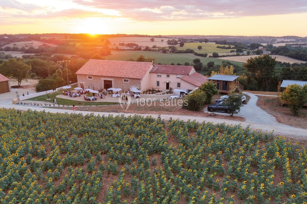 Vue aérienne d'une ferme entourée de champs de tournesols au coucher du soleil, avec des invités rassemblés à l'extérieur.