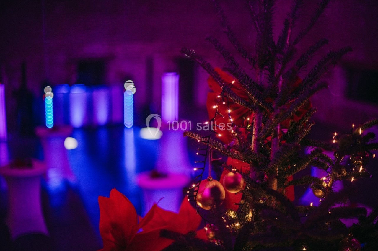 Sapin décoré avec guirlandes lumineuses et boules rouges, en arrière-plan une salle éclairée de lumières violettes.