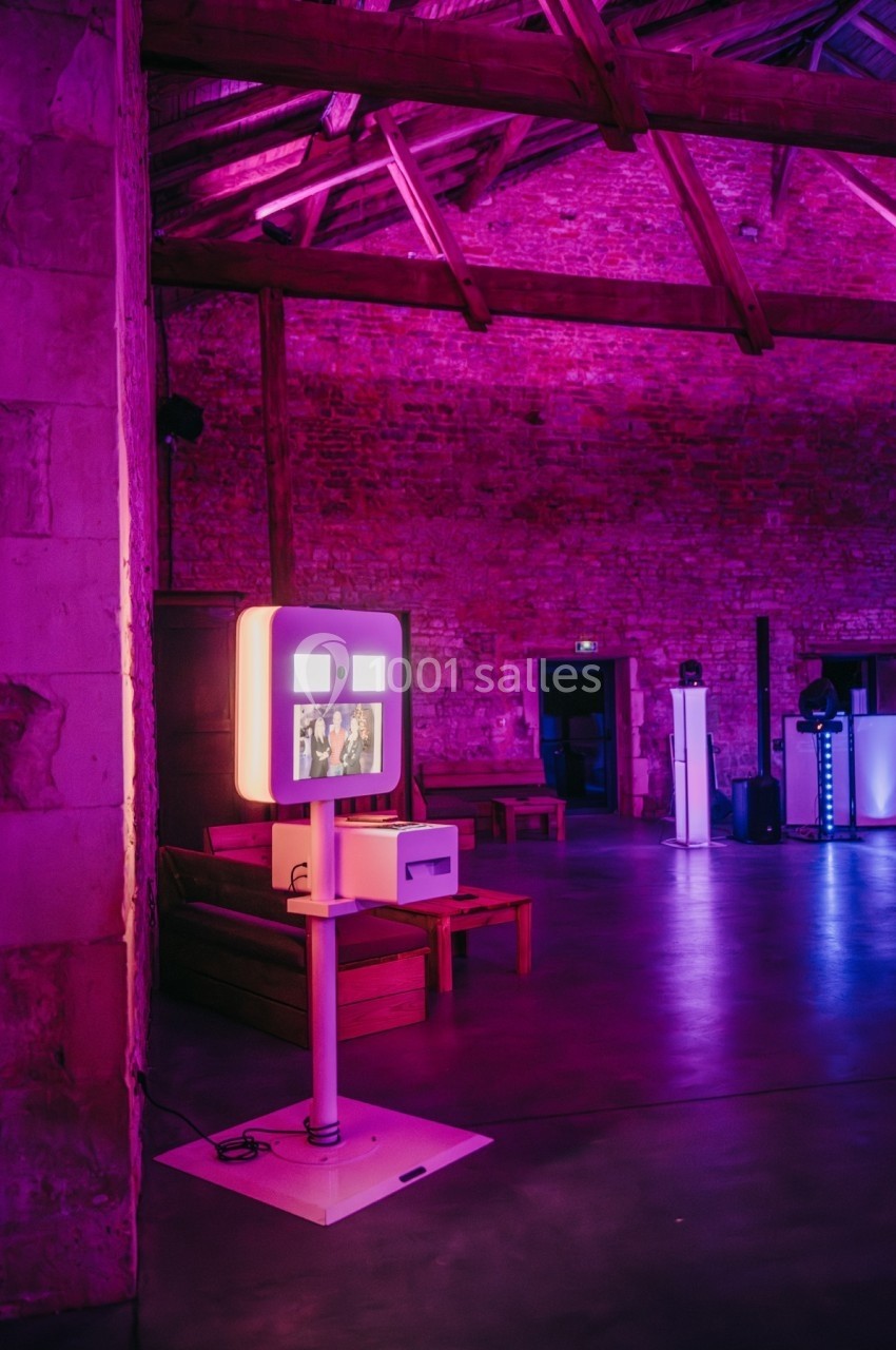 Borne photo interactive éclairée dans une salle en pierre avec éclairage violet et plafond en poutres apparentes.