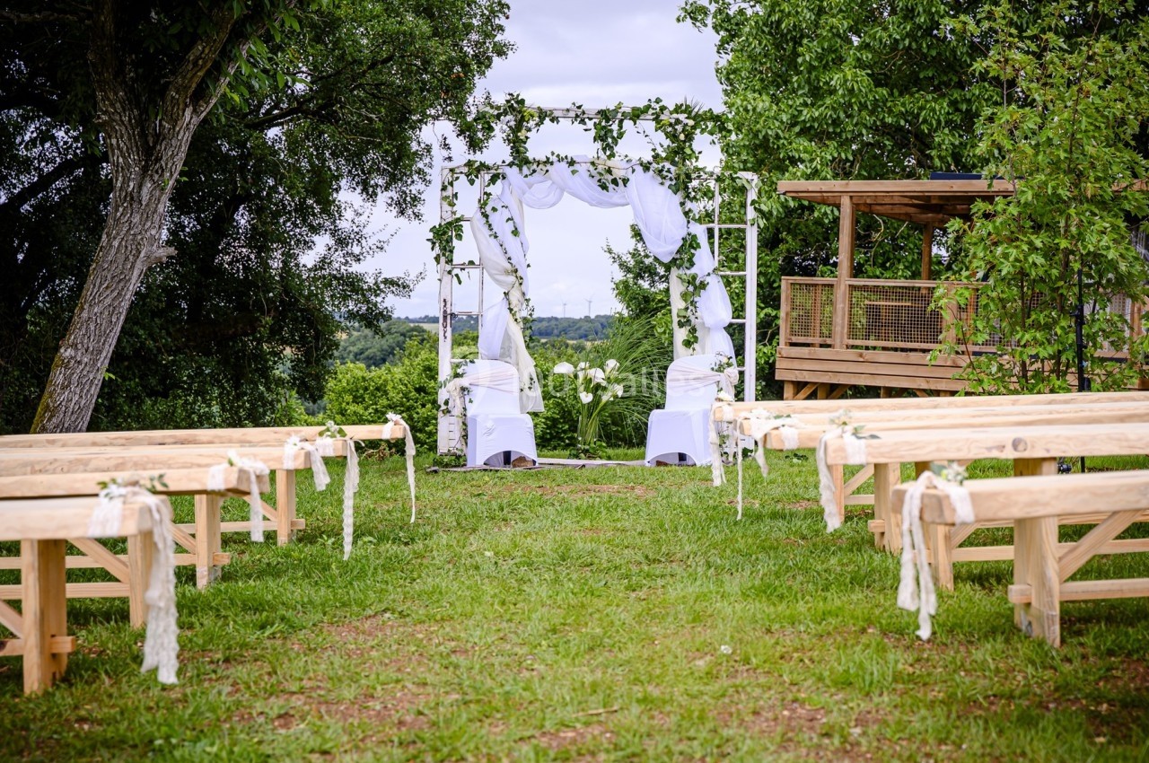 Allée extérieure avec bancs en bois menant à une arche décorée de tissus blancs et de feuillage pour une cérémonie.