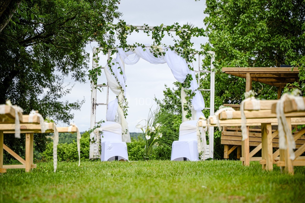 Arche de mariage décorée de voilages blancs et de feuillage, entourée de bancs en bois dans un cadre verdoyant.
