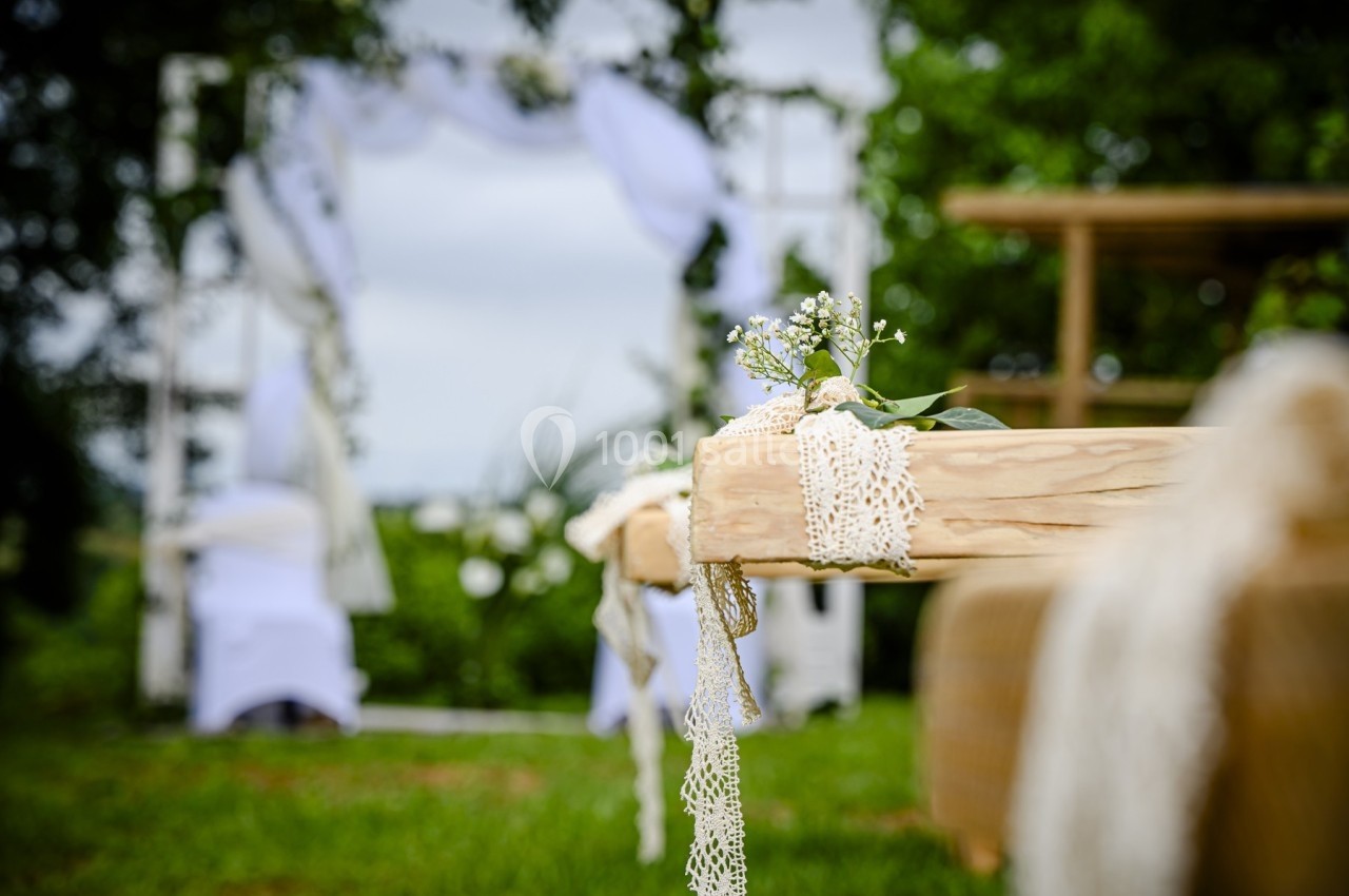 Banc en bois décoré de dentelle et de fleurs blanches, avec une arche de mariage floue en arrière-plan.
