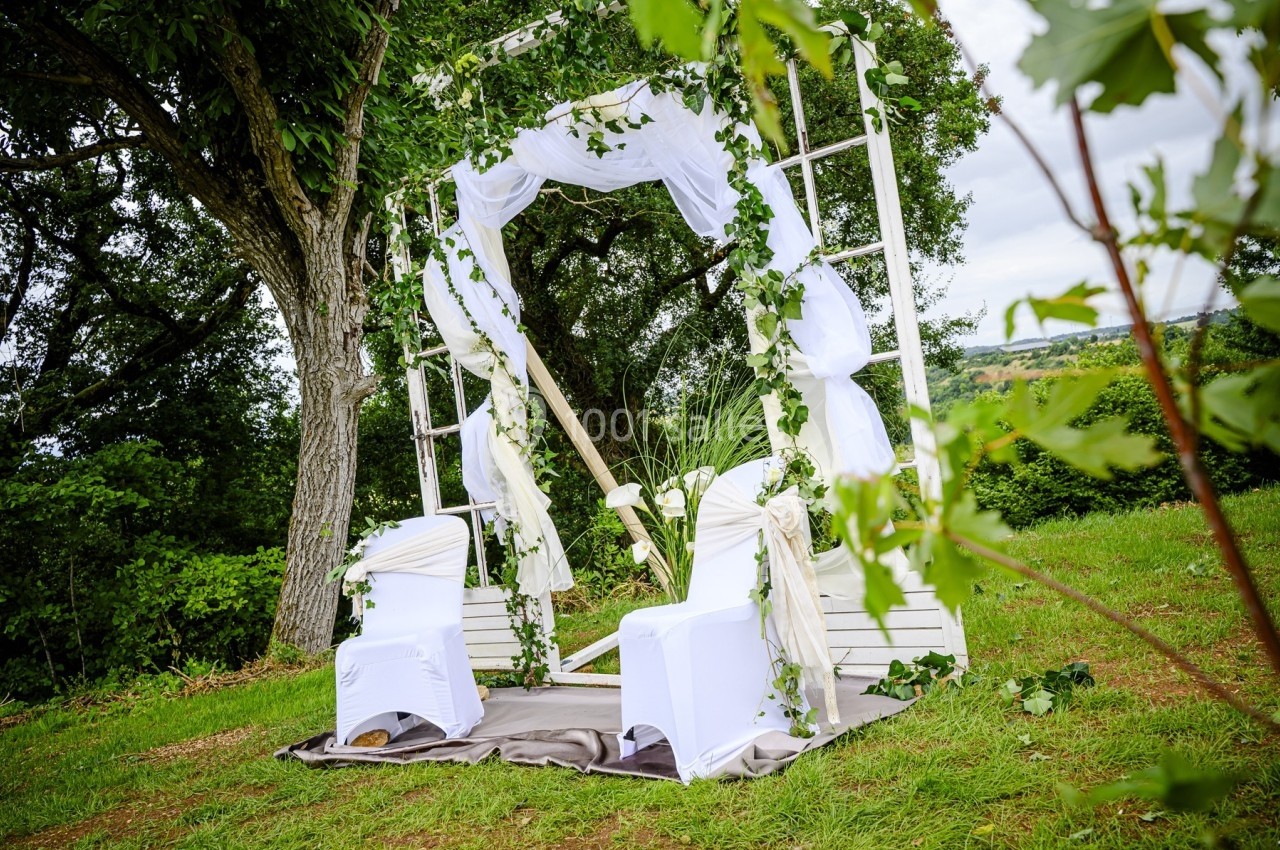 Arche de mariage décorée de voilages blancs et de feuillage, entourée de chaises blanches sur une pelouse verdoyante.