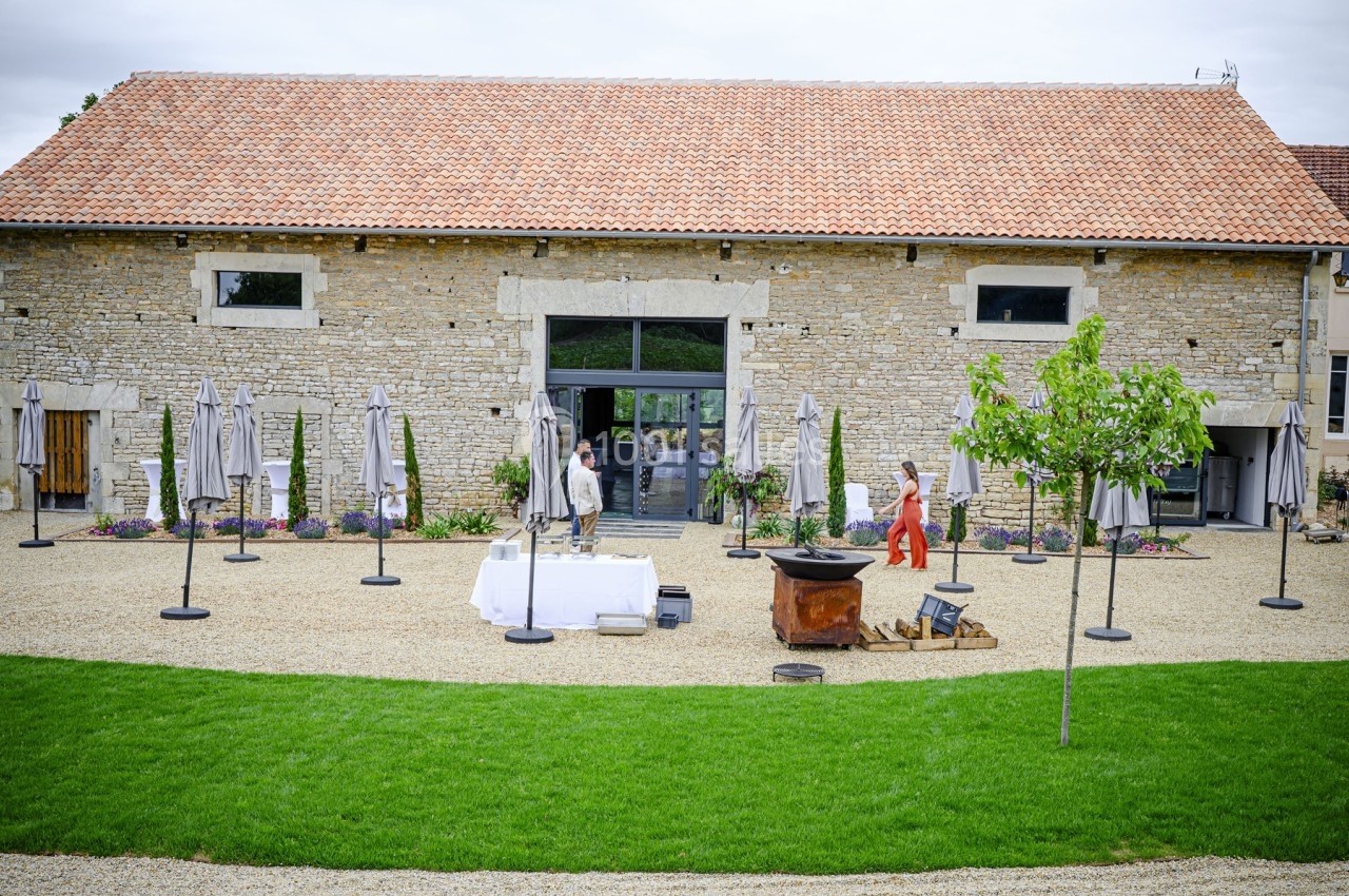 Grande bâtisse en pierre avec toit en tuiles, terrasse aménagée et deux personnes marchant dans la cour.