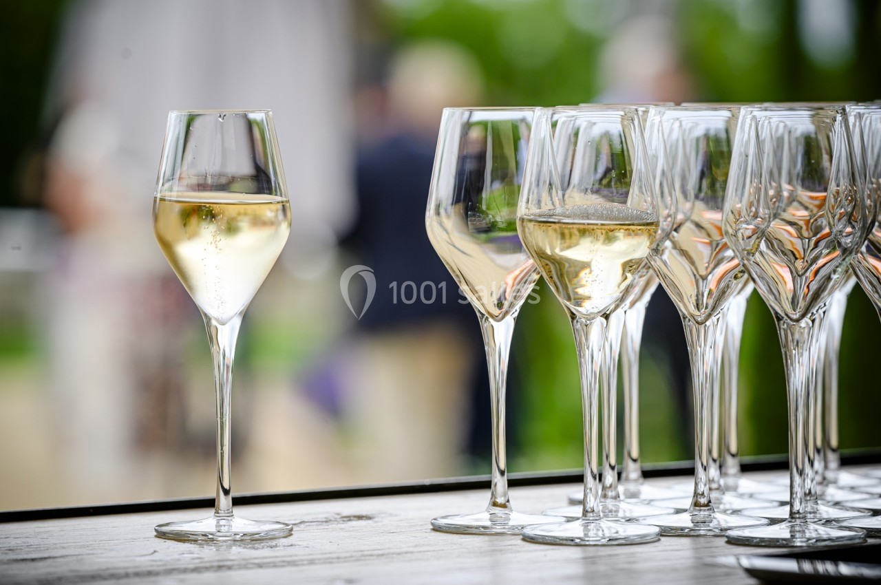 Verres de champagne alignés sur une table en extérieur, avec un arrière-plan flou de verdure et de silhouettes.