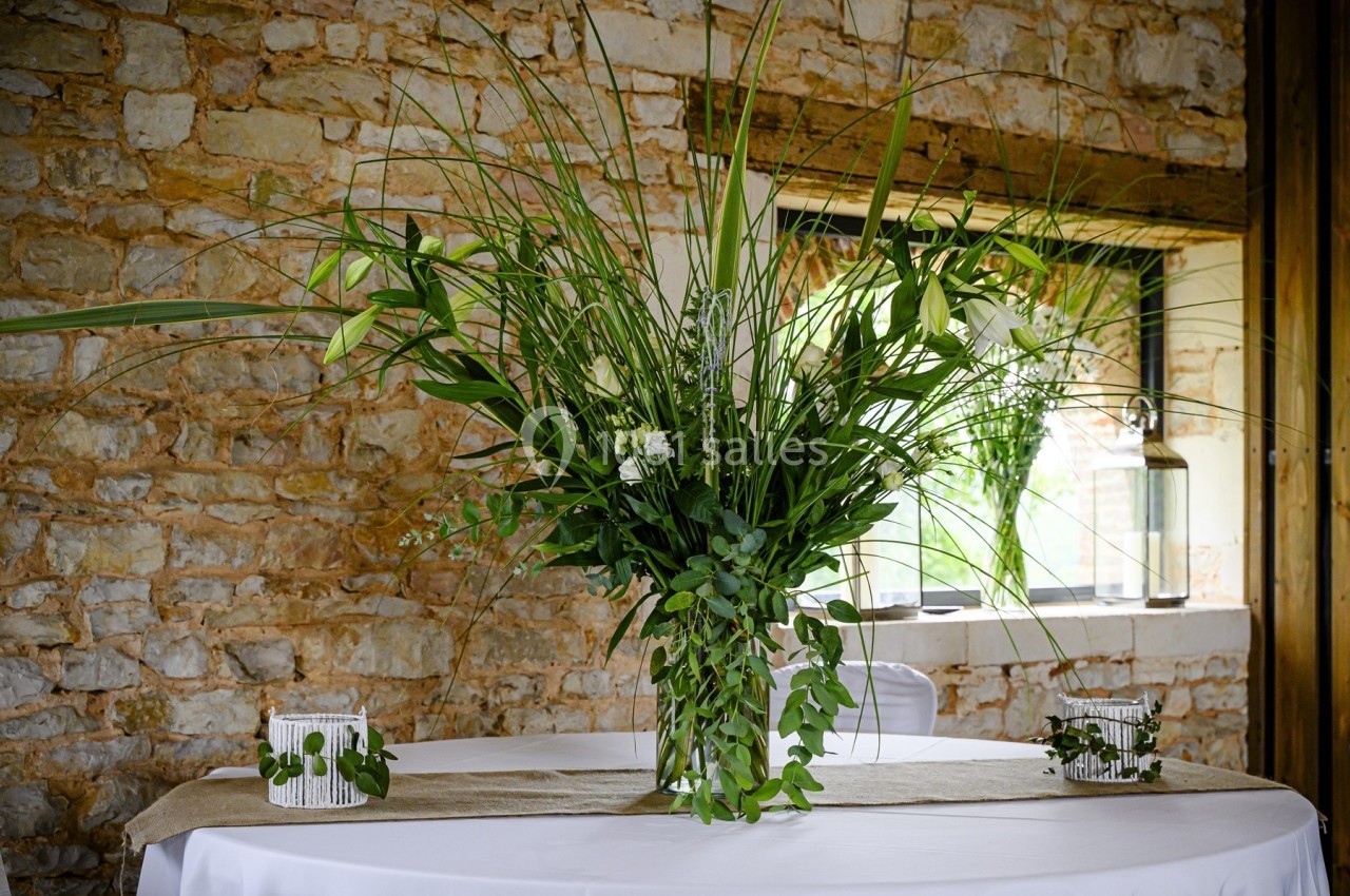 Arrangement floral vert et blanc sur une table ronde, devant un mur en pierre et une fenêtre lumineuse.