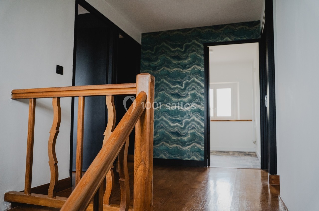 Vue d'un couloir intérieur avec un escalier en bois, un mur décoré de motifs ondulés et une pièce lumineuse au fond.