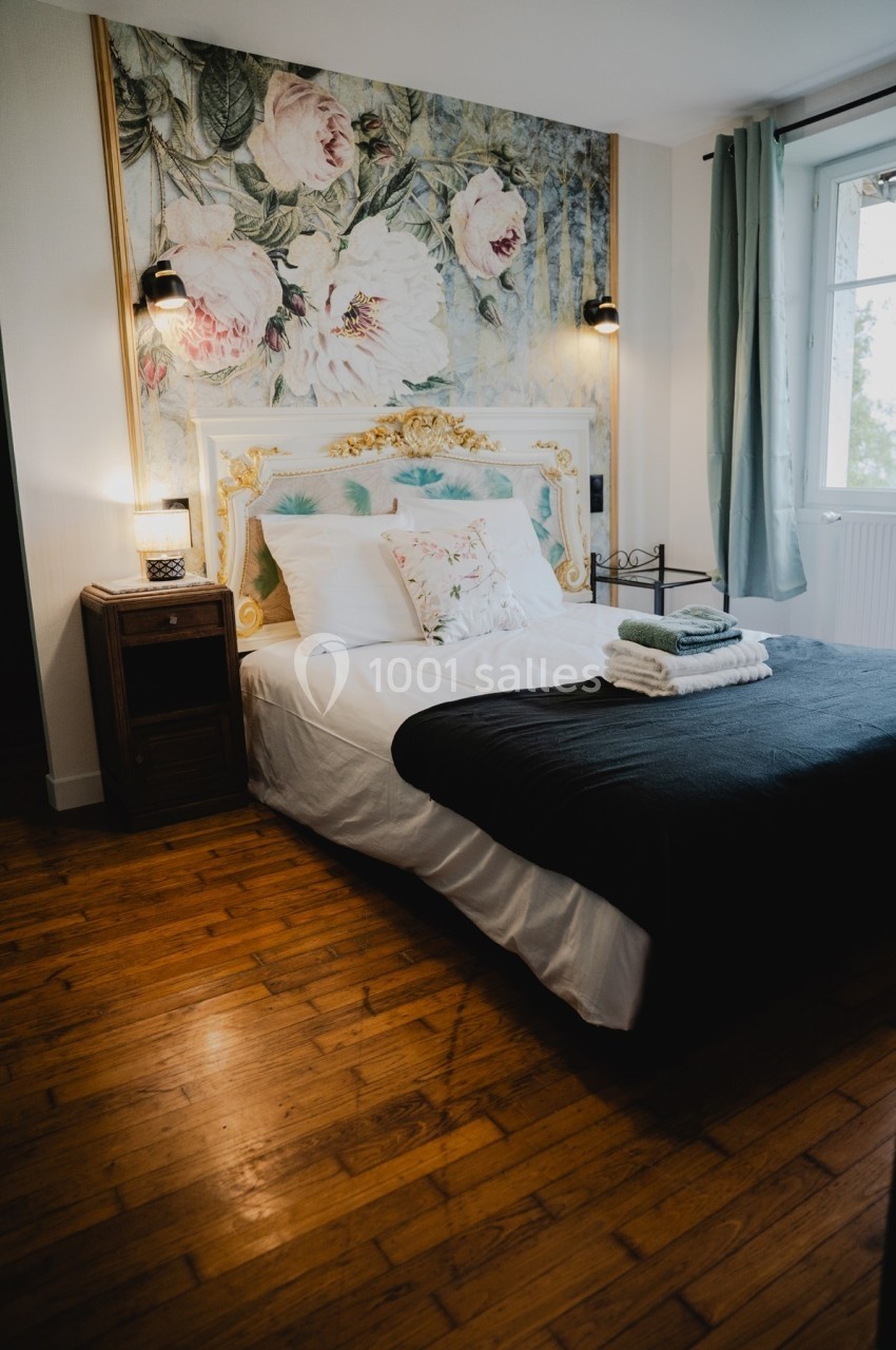 Lit double avec tête de lit ornée de motifs floraux, draps blancs, plaid sombre, et serviettes pliées sur un parquet en bois.